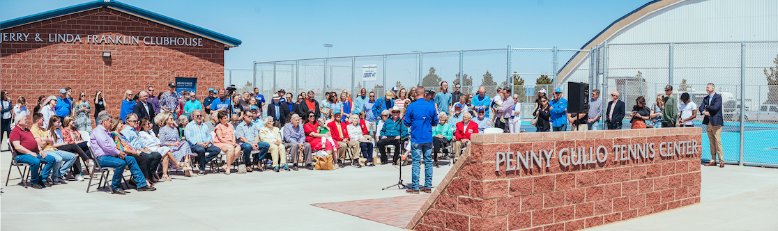 a crowd of onlookers watches a man giving a presentation in front of a newly-constructed tennis center and clubhouse