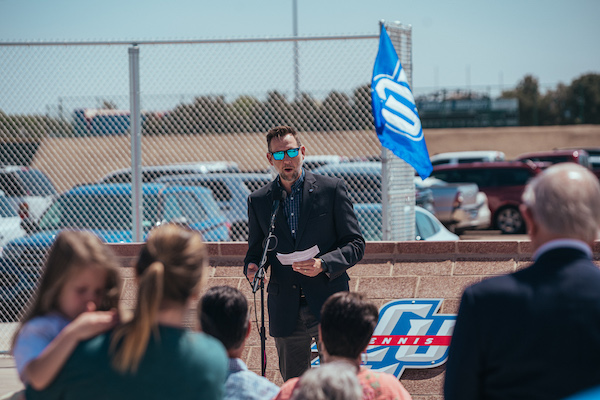 Jason Speegle, wearing a suit, stands at a microphone addressing an audience at the dedication ceremony
