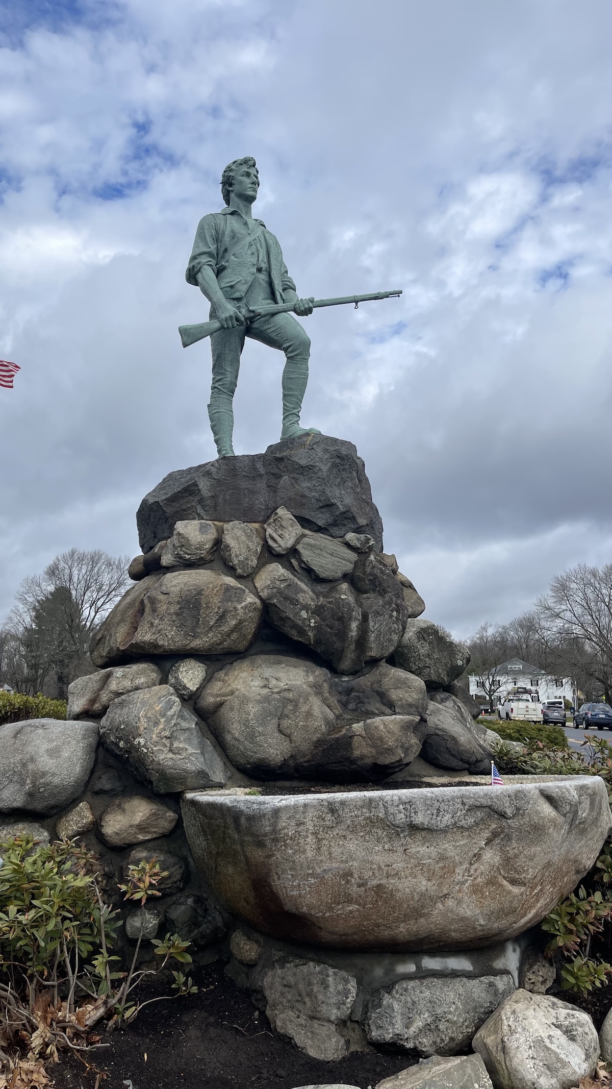 A tarnished copper statue of a colonial young man holding a musket loosely in front of him stands atop a mound of large stones.