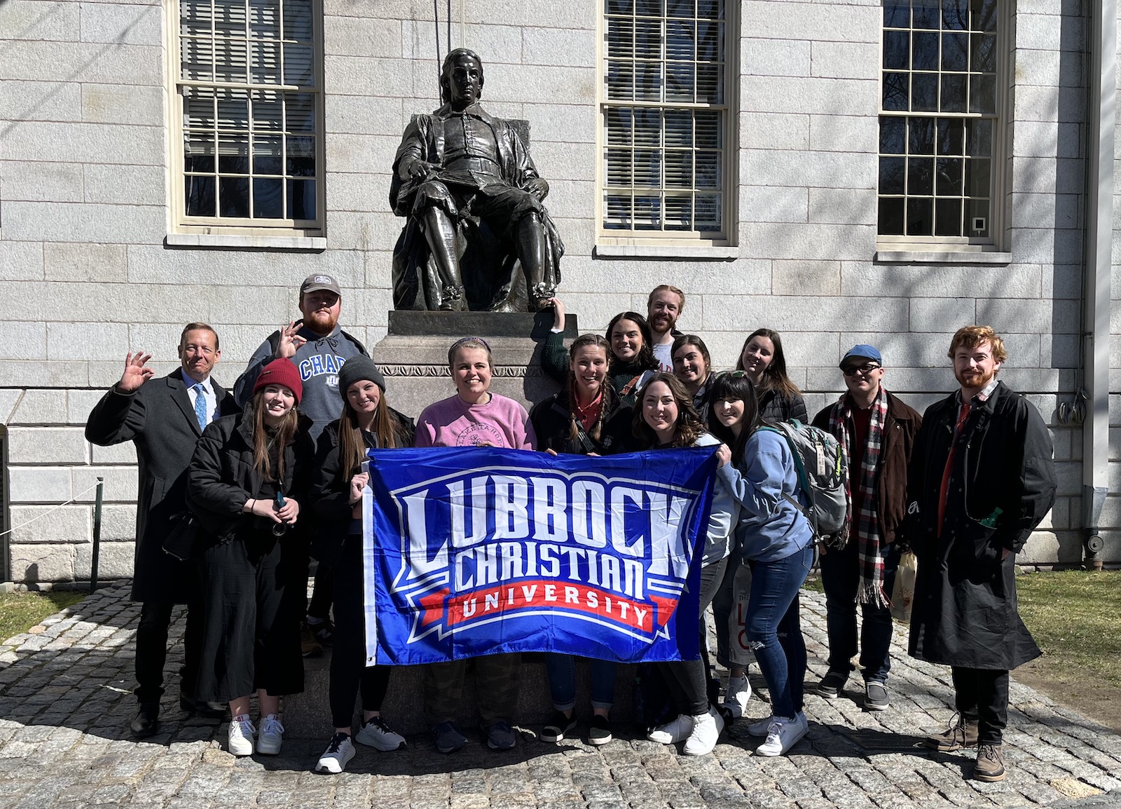 LCU students holding an LCU flag posing in front of a metal statue of John Harvard.