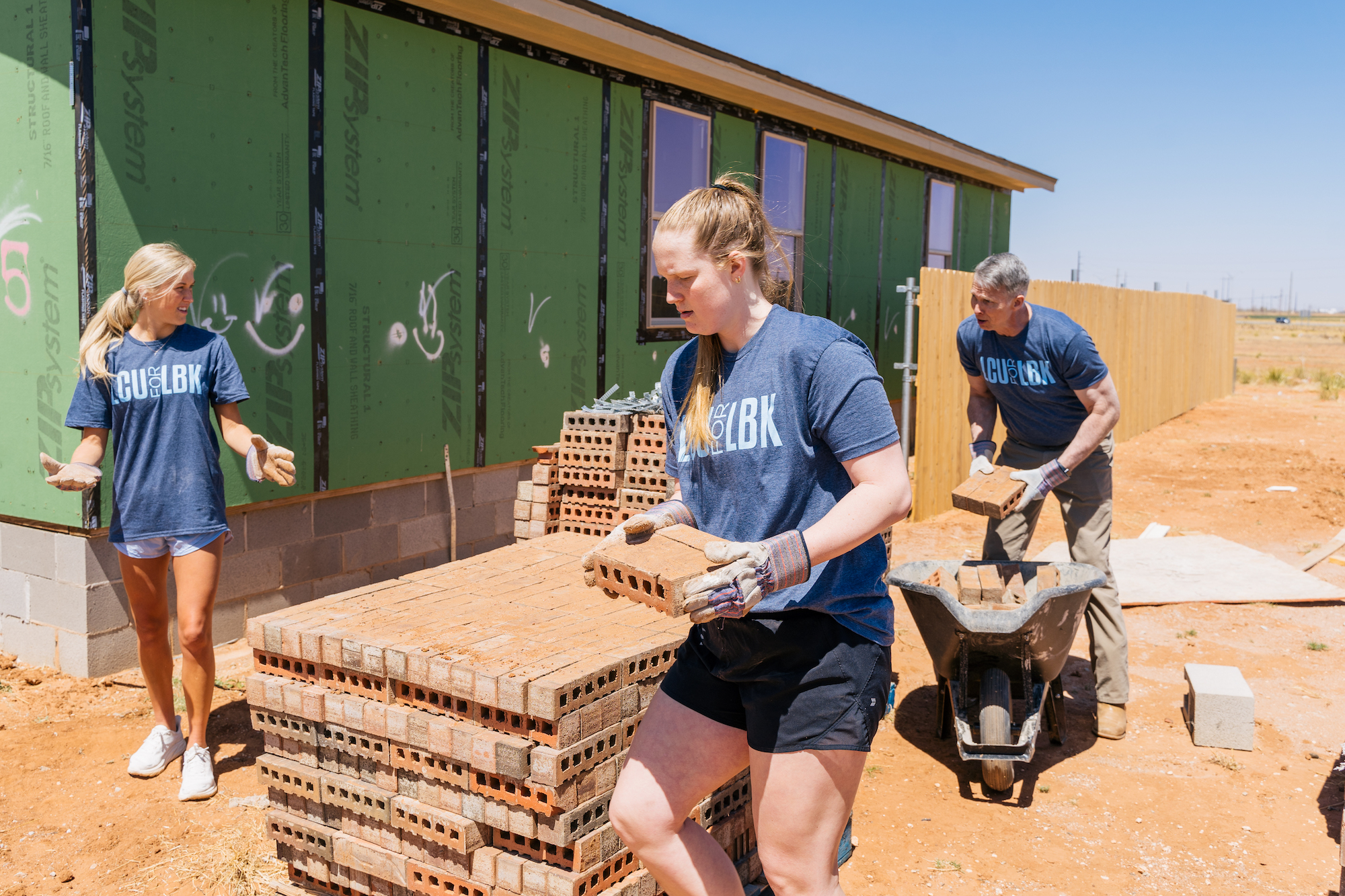 two young women carry bricks at a construction site, while an older gentleman places more bricks into a wheelbarrow behind them