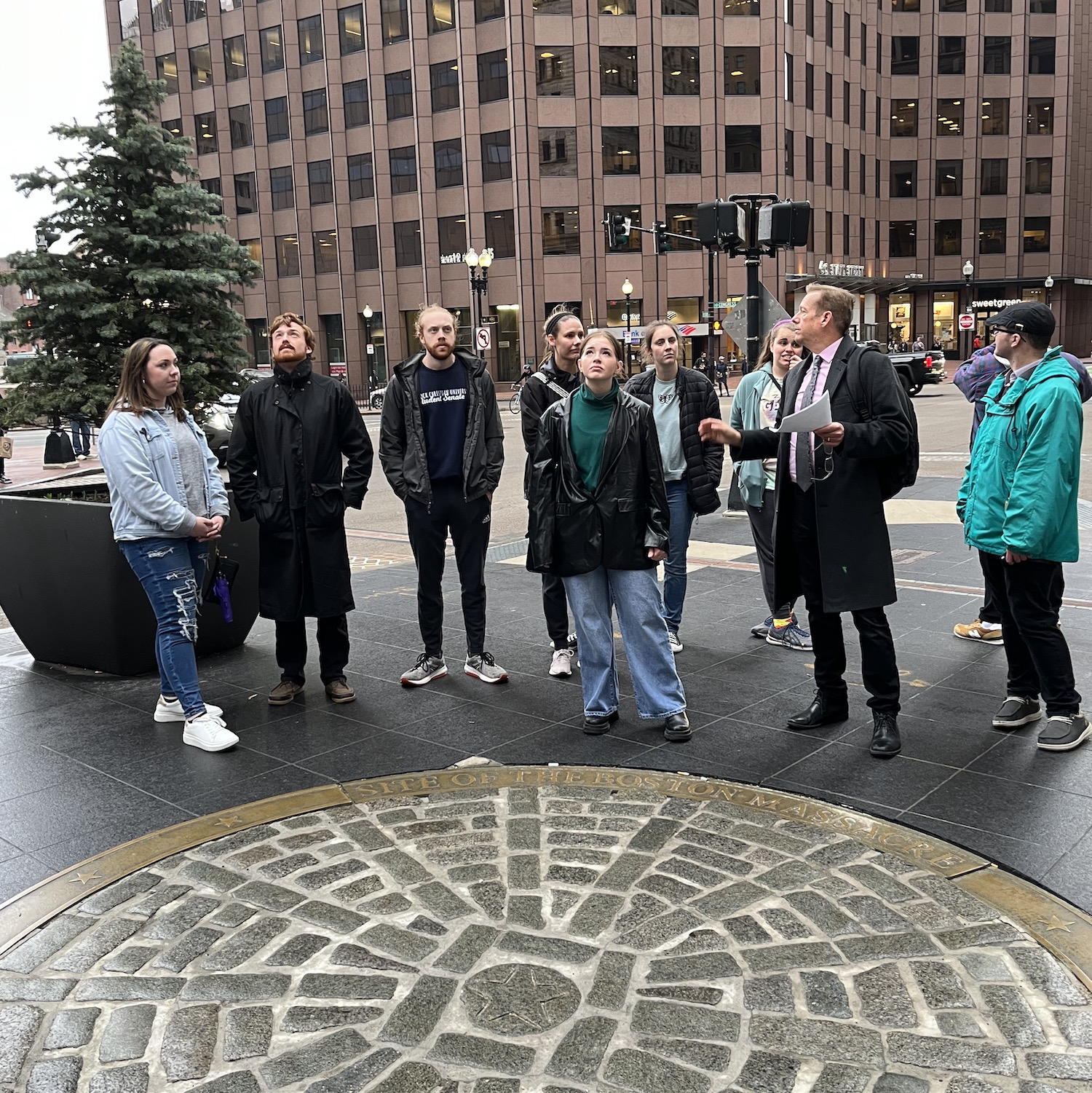 LCU students gather around a cobblestone circle in a city square, around which a plaque reads: Site of the Boston Massacre. The professor is giving a lecture.