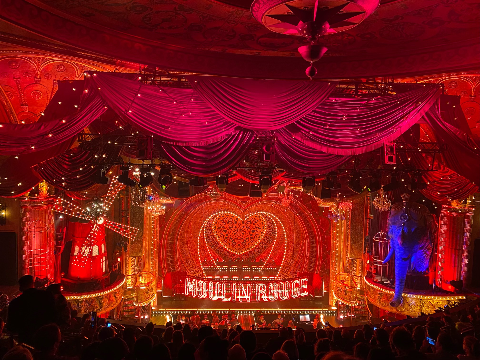 A photo looking down from the audience at an elaborately-decorated theatre stage. The set features gaudy hanging fabrics, many lights, and the words "Moulin Rouge" in large letters across the stage.