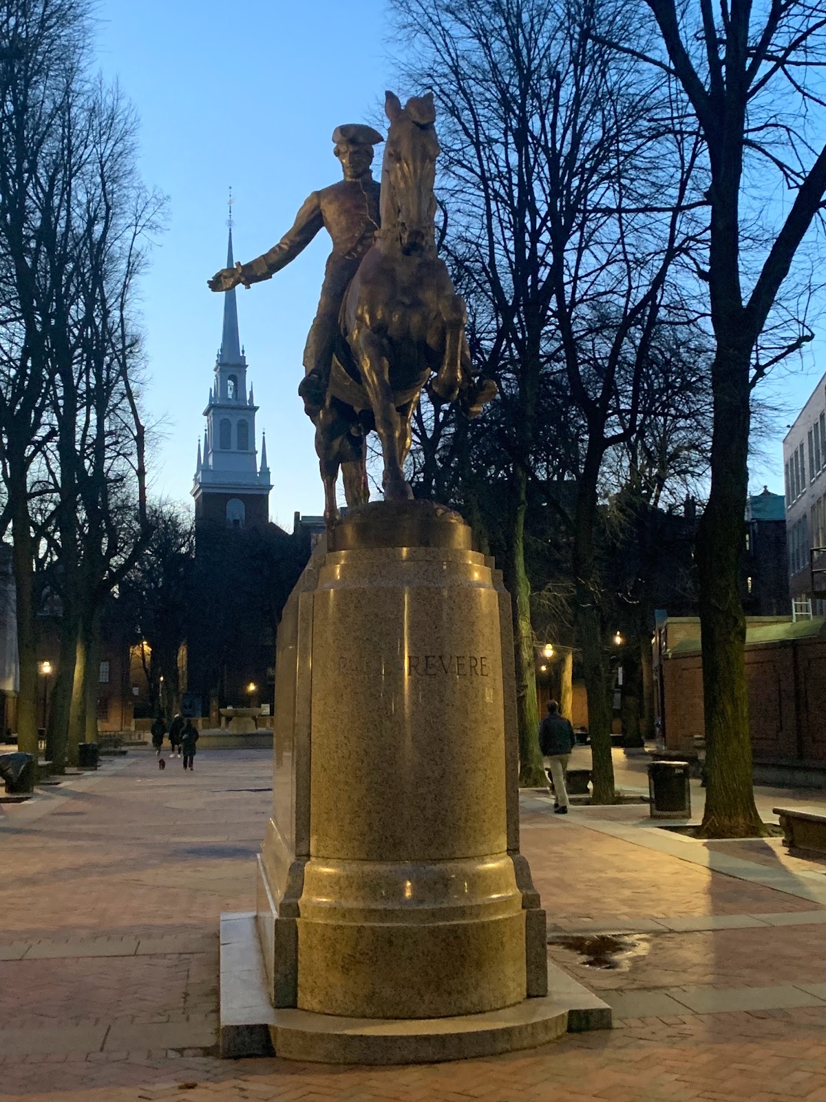 A metal statue featuring a man riding on a horse on top of a pedestal with the name Paul Revere inscribed in the stone. In the background, a historic church looms.