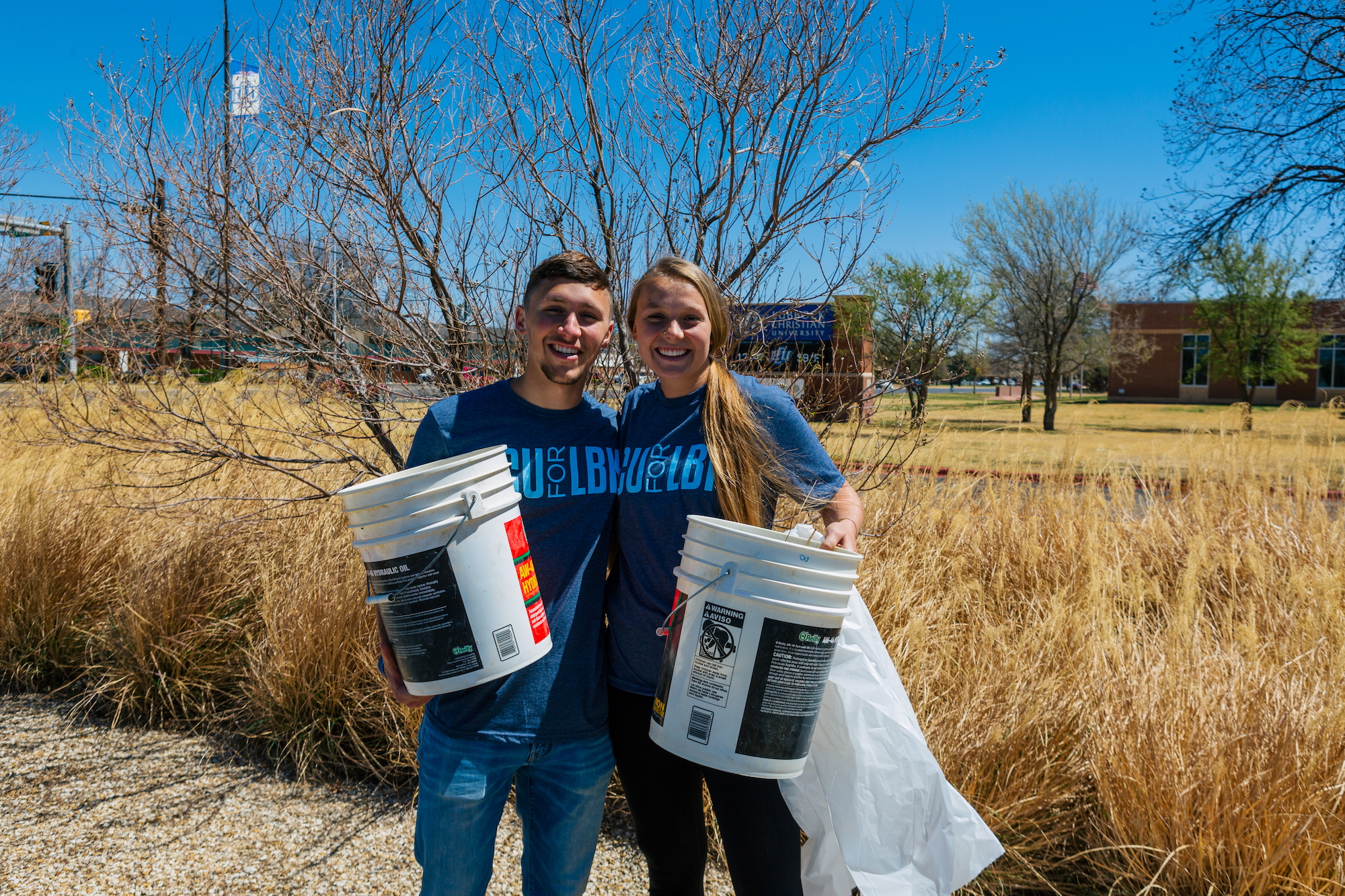 two students holding buckets for picking up trash smiling beside foliage