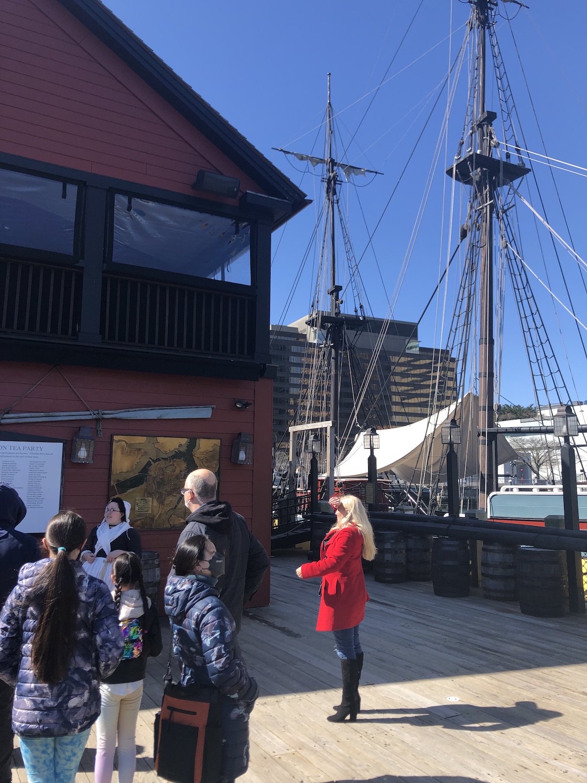 Tourists standing in front of the Tea Party museum. In the background, the rigging of a colonial ship can be seen next to a dock.