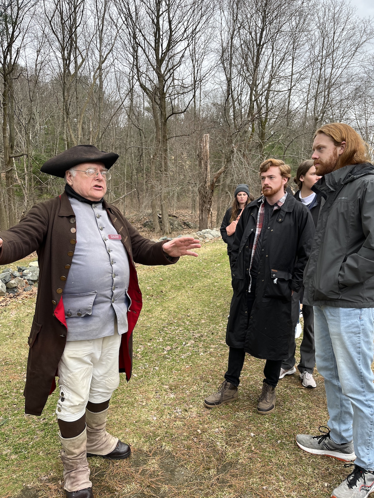 a man dressed in American colonial garb speaks to students outside near a copse of trees