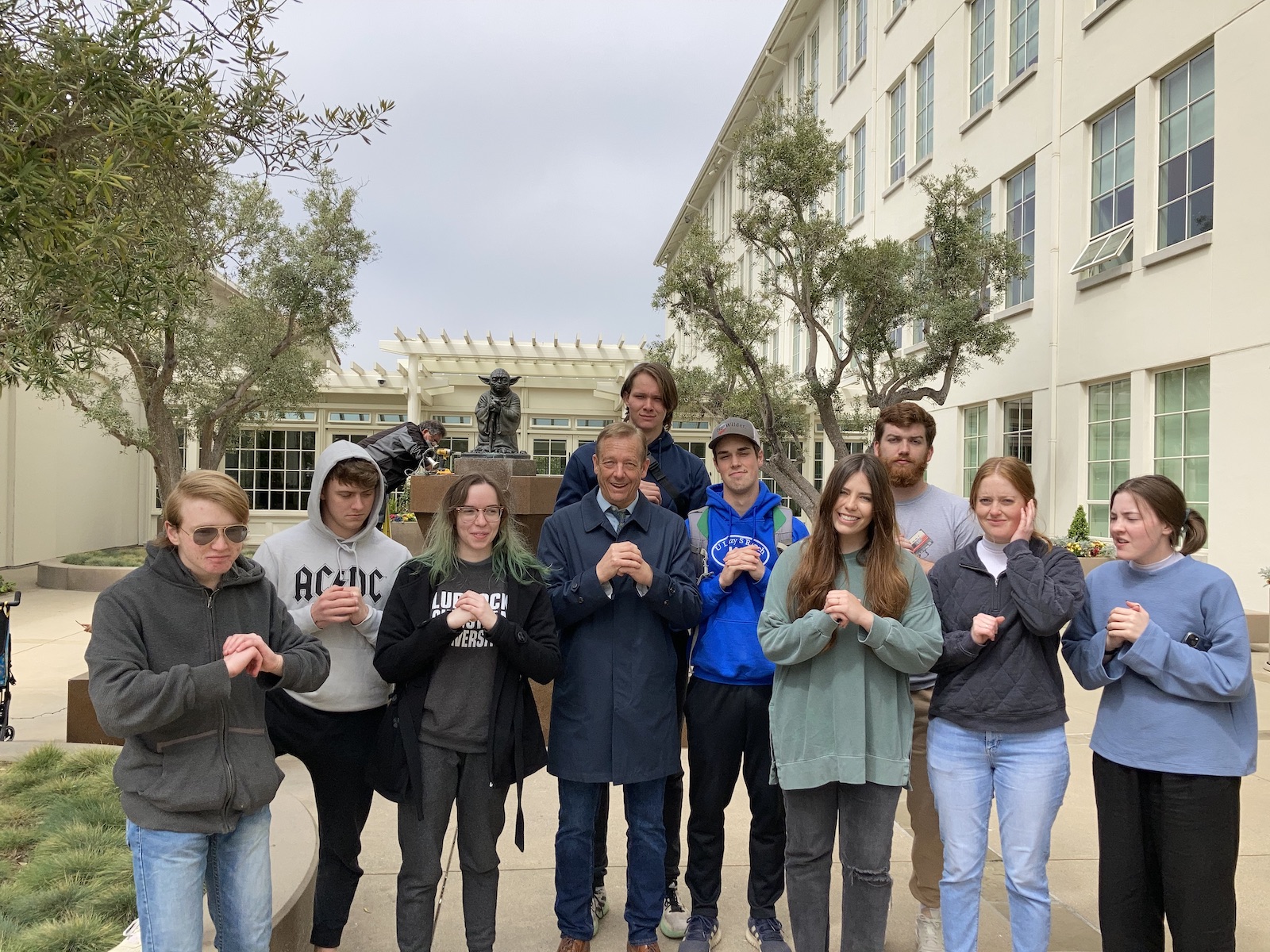 Dr. Fehr and his class pose with hands mimicking Yoda leaning on his cane, in front of the statue of Yoda at Lucasfilm.