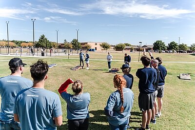 a group of people around a corn hole tournament in a grassy area of the plaza