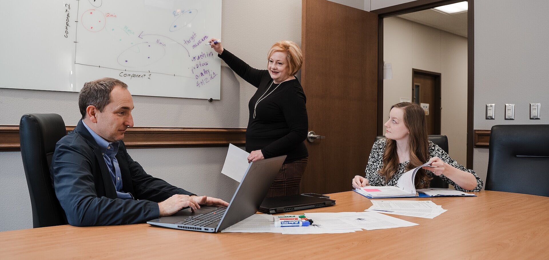 Dr. Julie writing on a white board while Drs. Fisher and Cherry sit at table analyzing data.