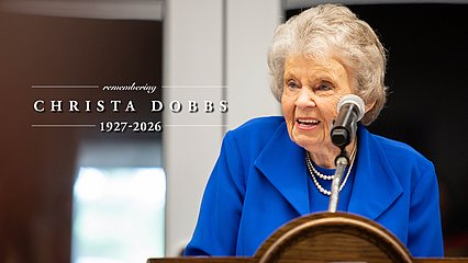 Christa Dobbs behind a lectern and the words, "Remember Christa Dobbs: 1927-2026"