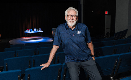 Don standing behind the chairs of the theatre and lit stage 