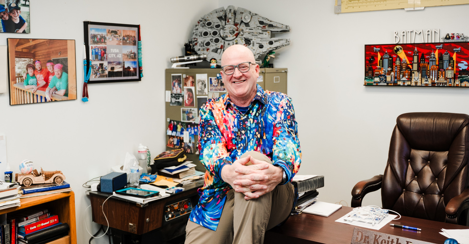 Keith sitting on his desk in his office with a colorful shirt on